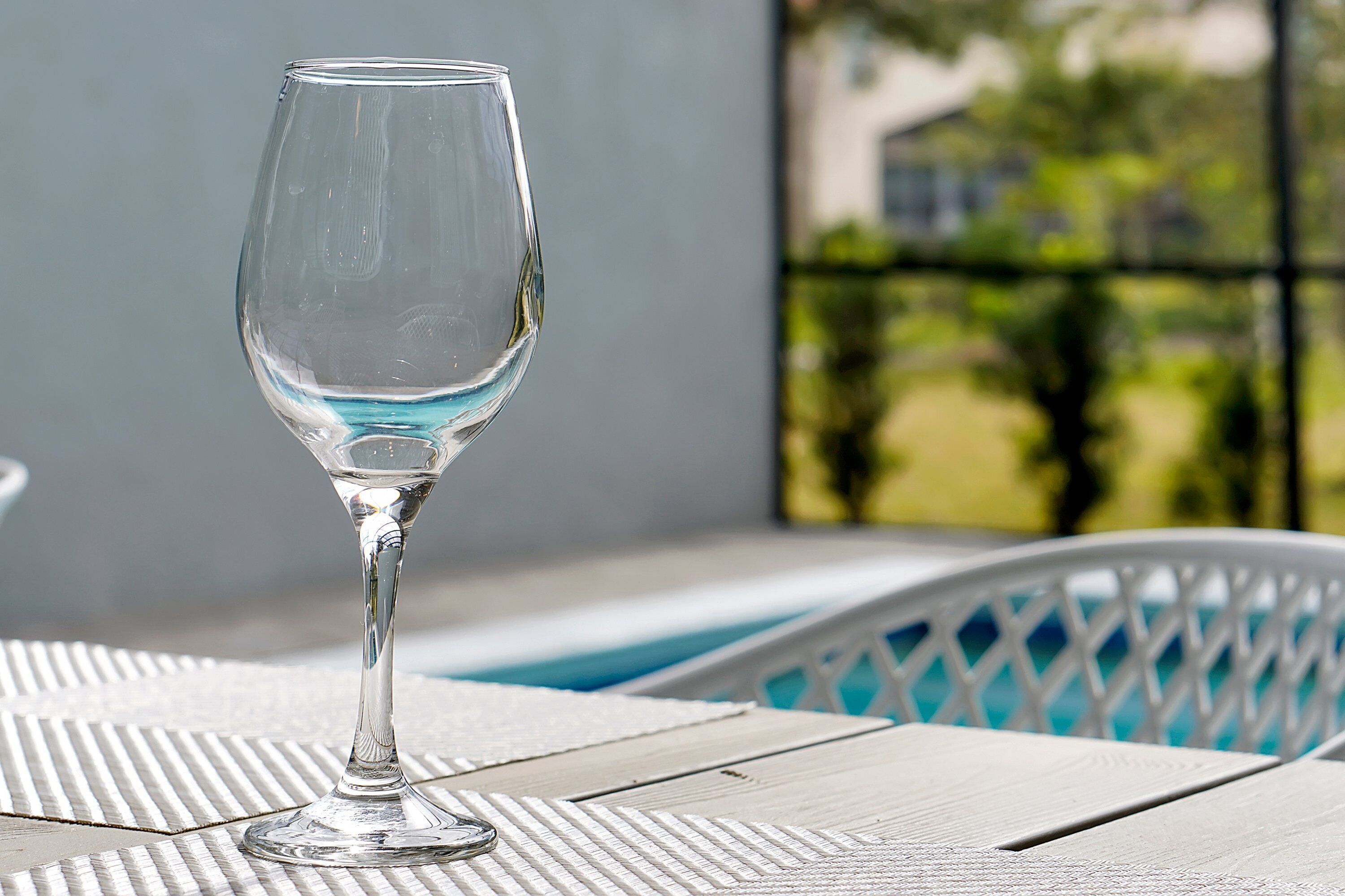 A clear glass wine glass on a textured table with a view of a pool and greenery in the background.