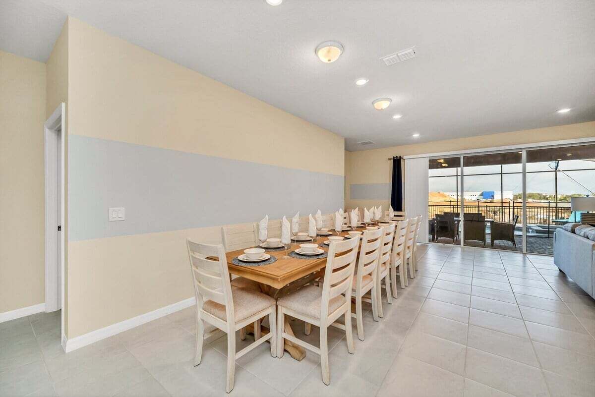 A dining area with a long table set for six, surrounded by chairs, in a room with a large window and a view of the outside.