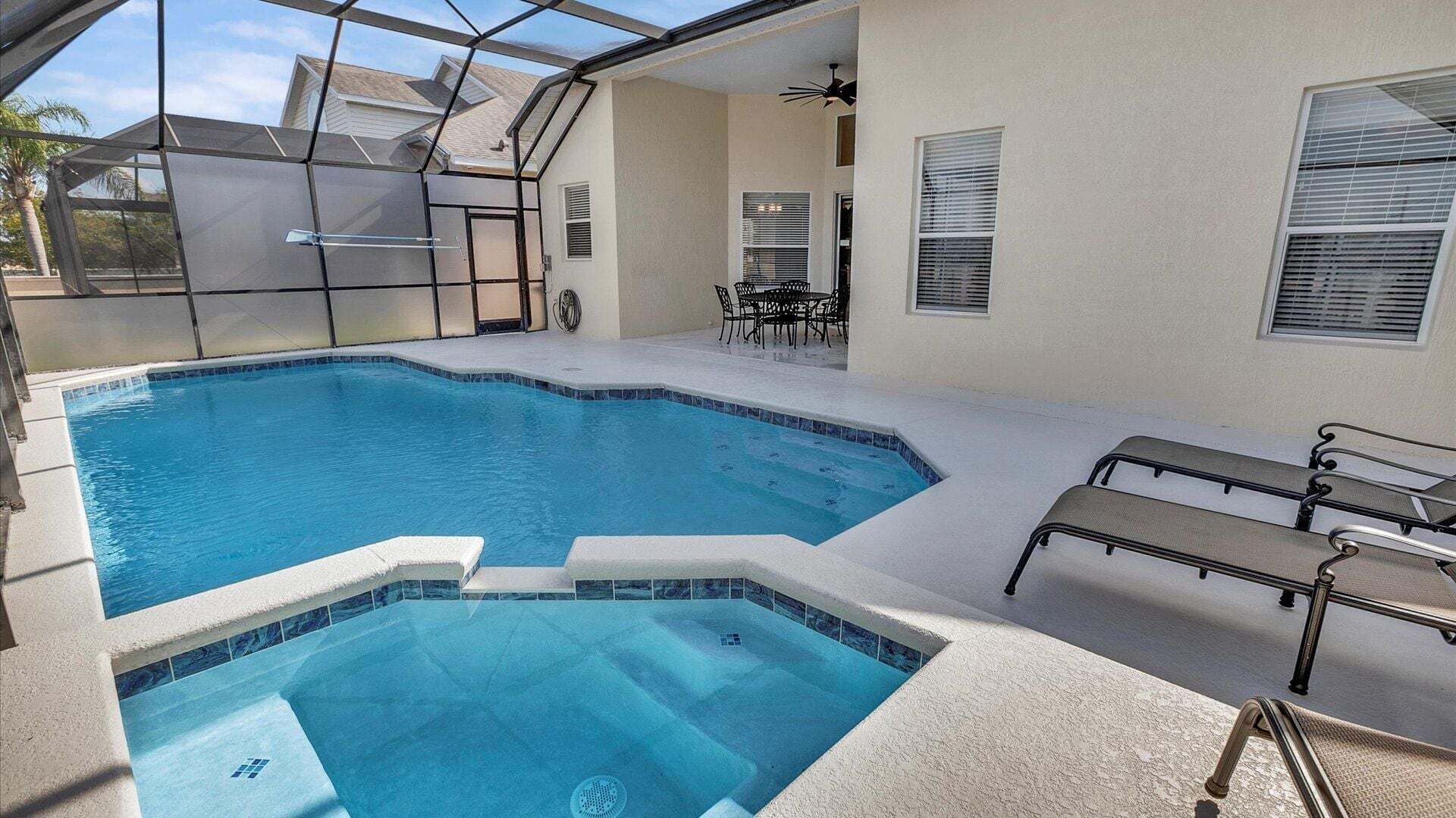 A swimming pool with a glass enclosure, a hot tub, and a patio area with chairs.