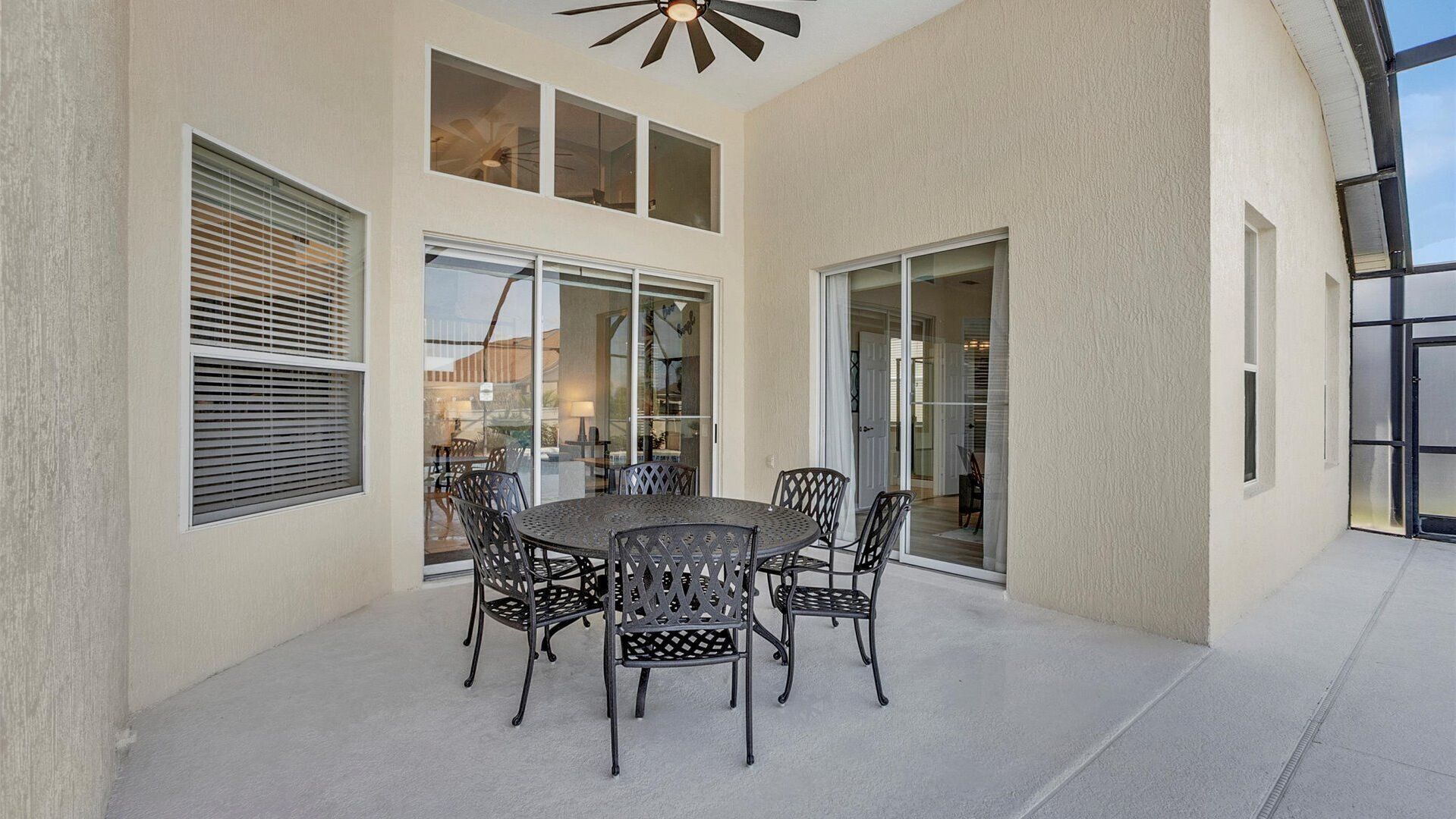 An outdoor patio with a round table and chairs, large windows, and a ceiling fan.