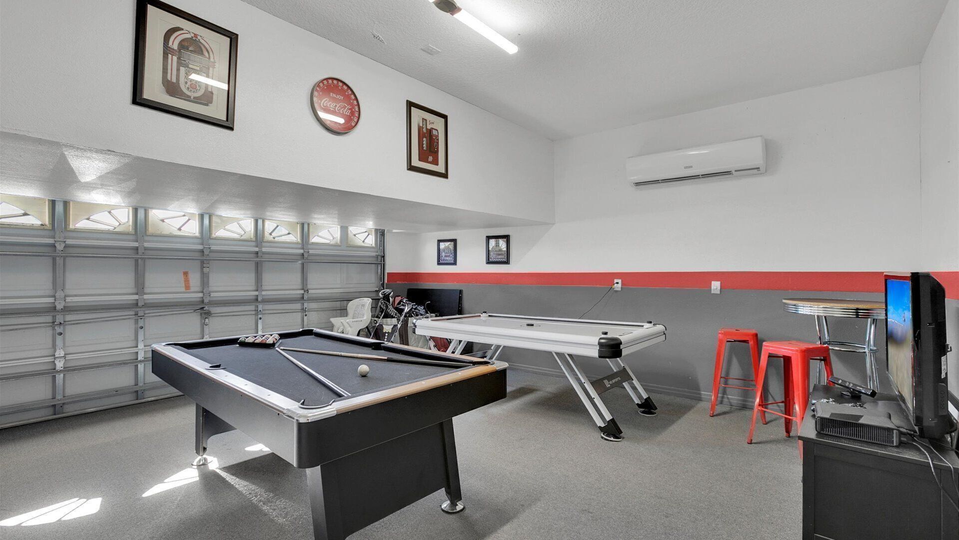 Een biljart in een ruimte met een garagedeur, twee ingelijste foto's en een airconditioning.