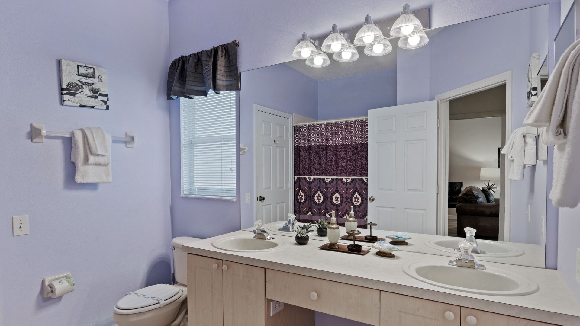 A bathroom with two sinks, a mirror, and a toilet. There are towels on a rack and a window with blinds.