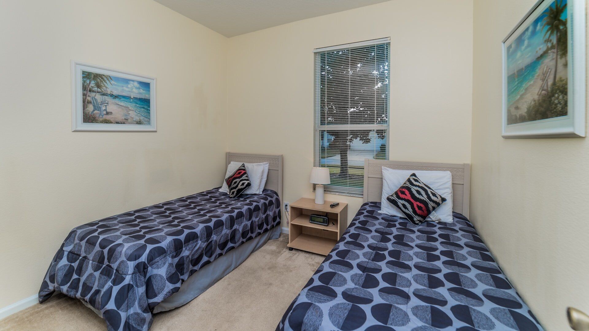 A room with two single beds, each with a patterned bedspread and matching pillows. There is a nightstand with a lamp and a small table between the beds. The room has a window with blinds, and there are two framed pictures on the walls.