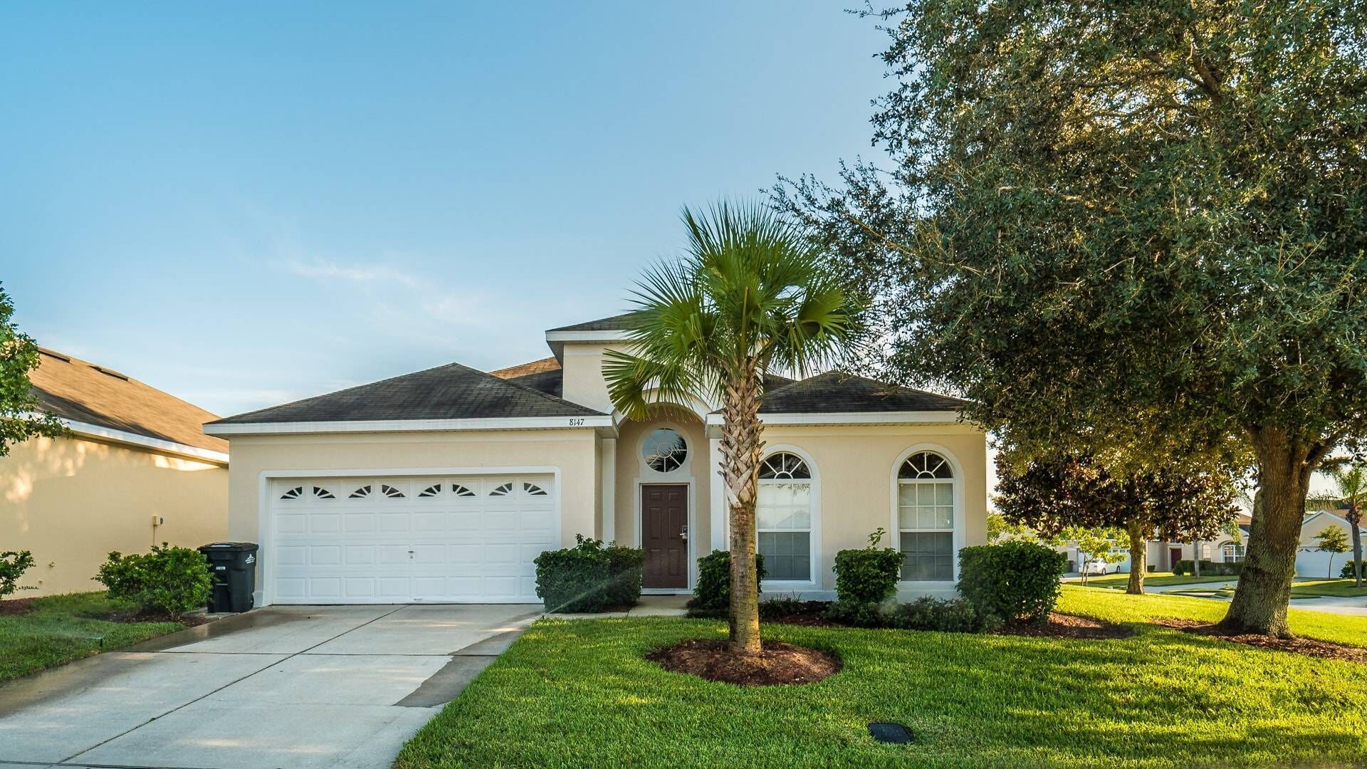 A single-story house with a garage, a front door, and a small front yard with a palm tree and shrubs.