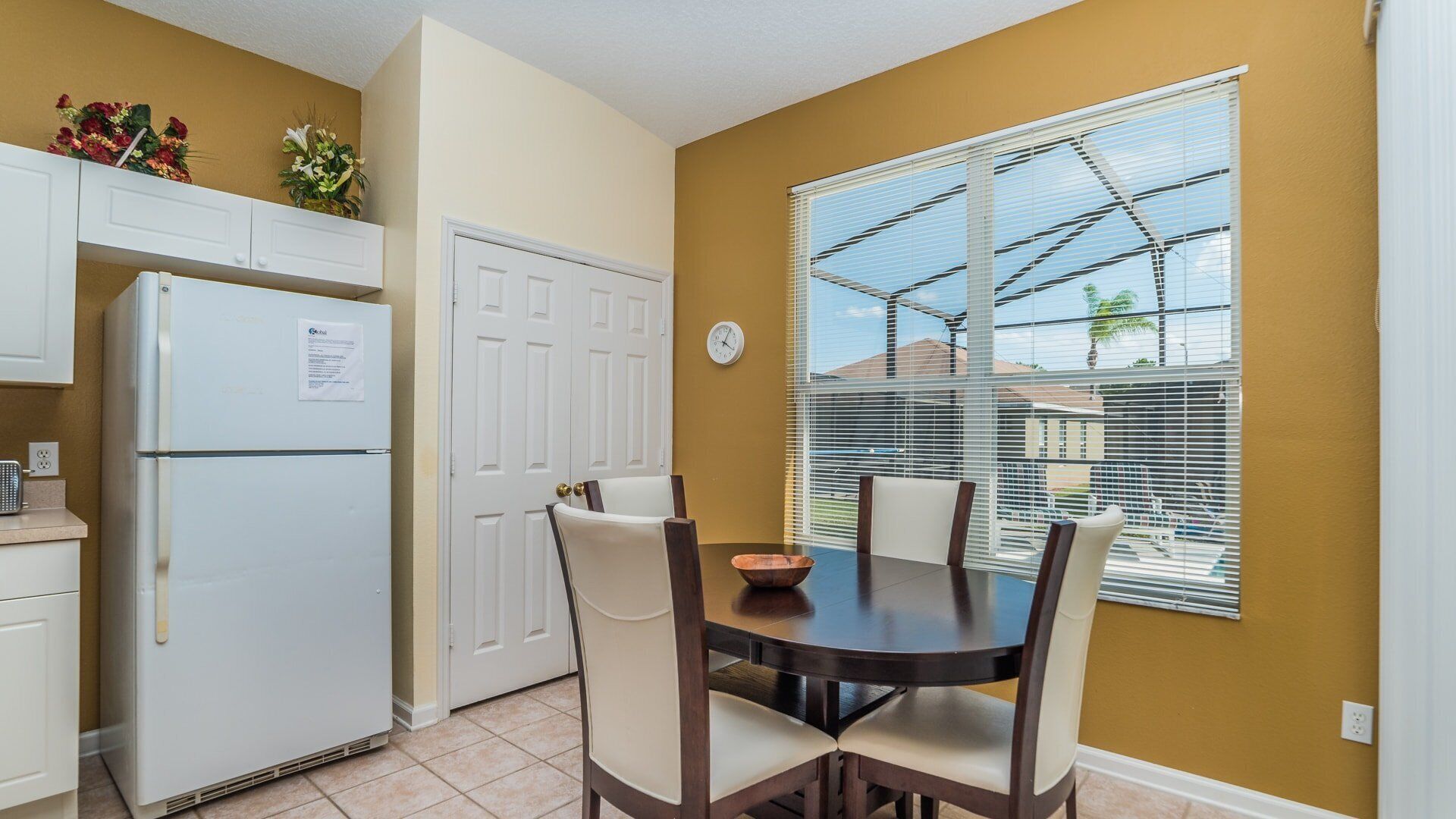 A compact kitchen with a refrigerator, a dining table with chairs, and a window with blinds.