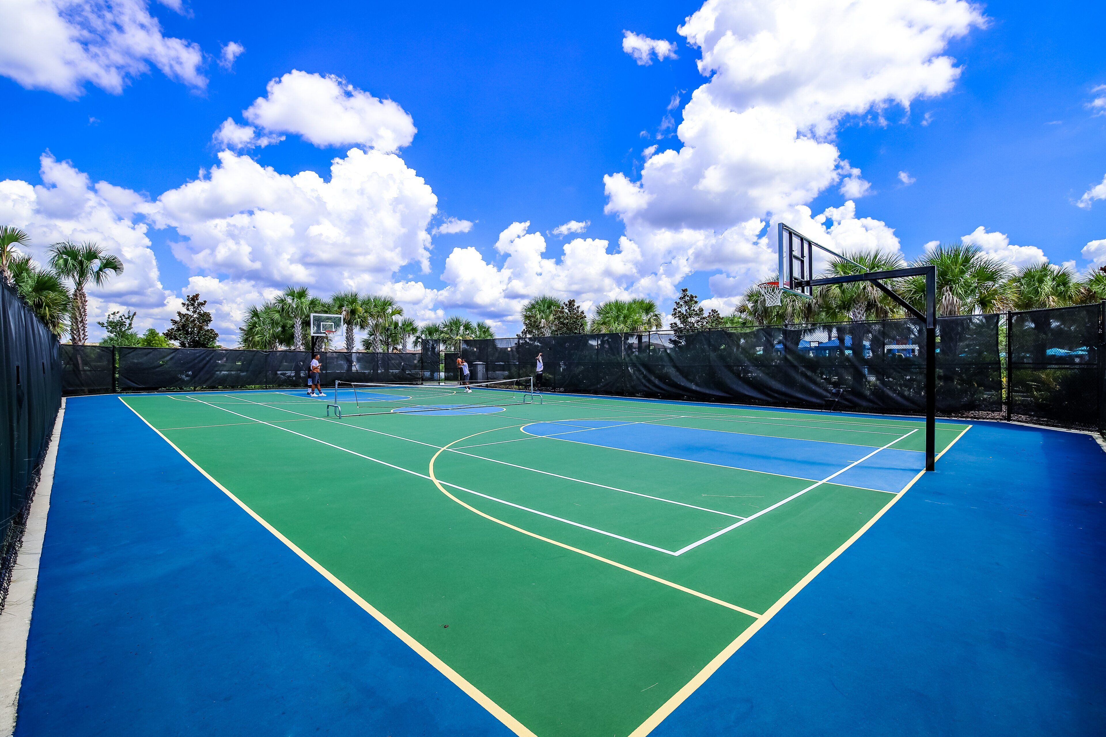 Tenniskenttä, jossa on myös koripallokehä, ja kentän ympärillä on palmuja sekä kirkas taivas.