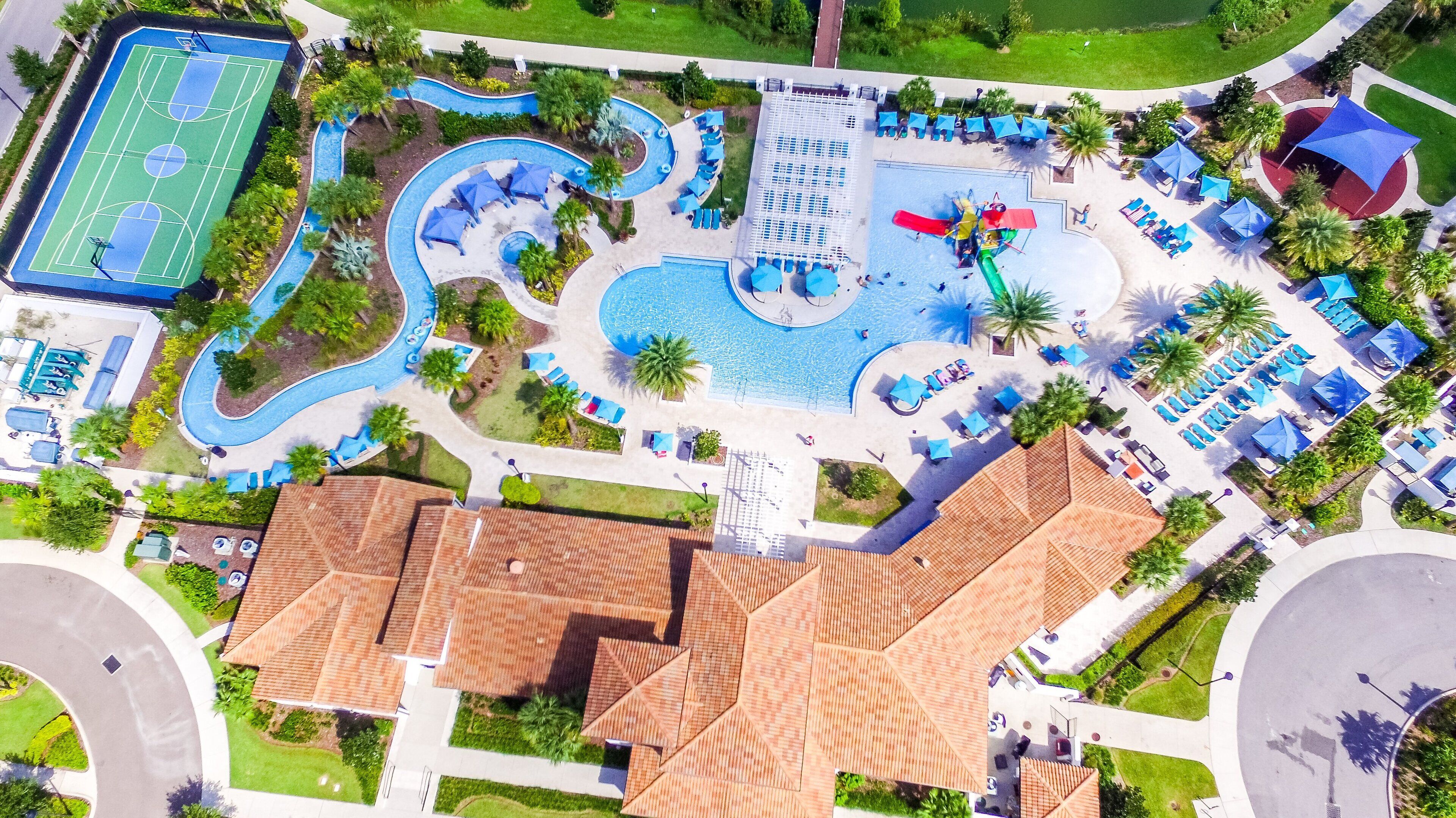 An aerial view of a resort with a swimming pool, sun loungers, a tennis court, and a water slide.
