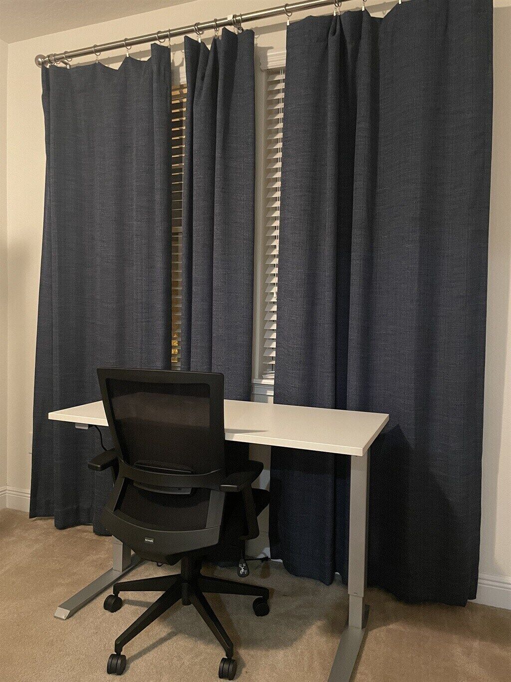 Habitació amb escriptori blanc, cadira d'oficina negra i cortines fosques.