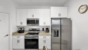 Una cocina compacta con armarios blancos, un refrigerador de acero inoxidable, un microondas, un horno y una cafetera.