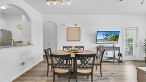 Un comedor con una mesa redonda, cuatro sillas, una cocina con un fregadero y grifo, un reloj y un televisor en la pared.