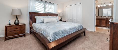 Quarto com cama de madeira, mesa de cabeceira, luminária, janela com cortinas, porta e banheiro visível através de uma porta aberta.