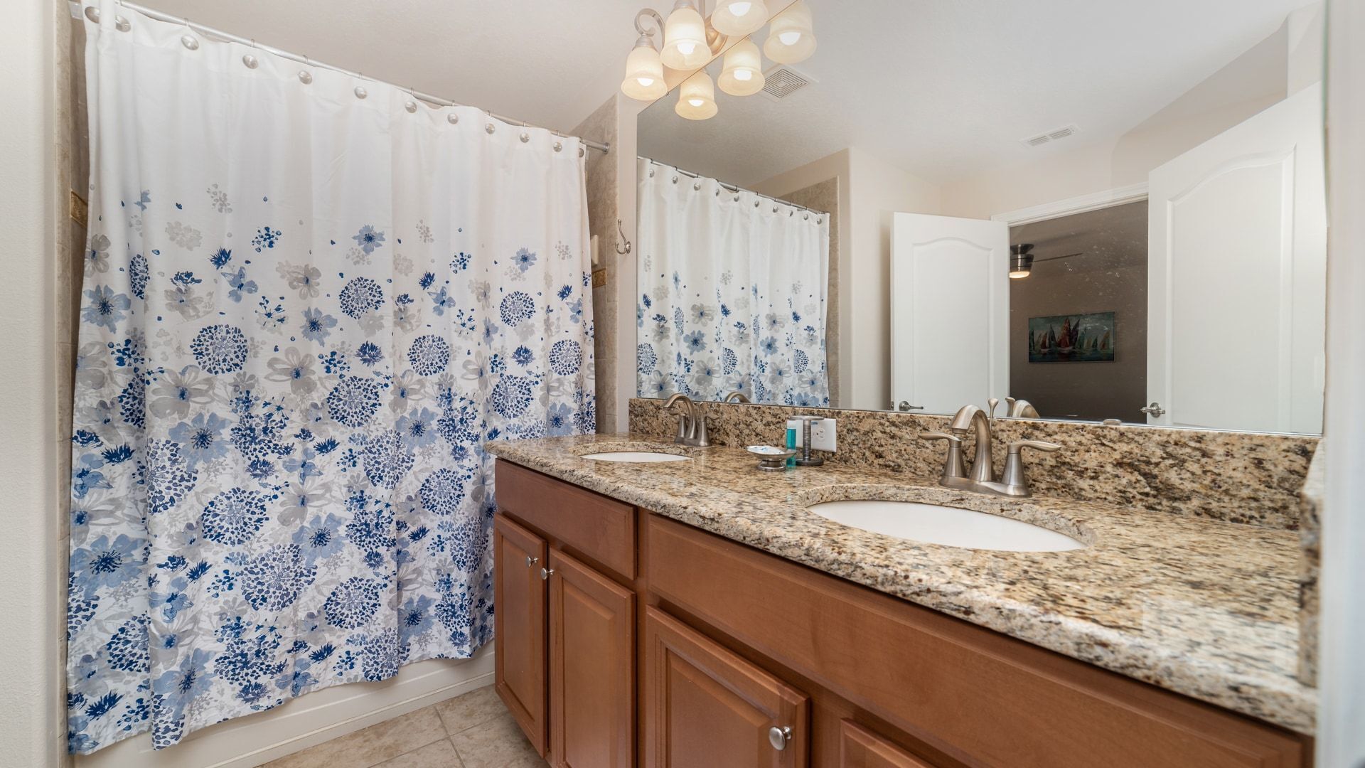 A bathroom with two sinks, a granite countertop, wooden cabinets, a large mirror, and a shower with blue floral curtains.