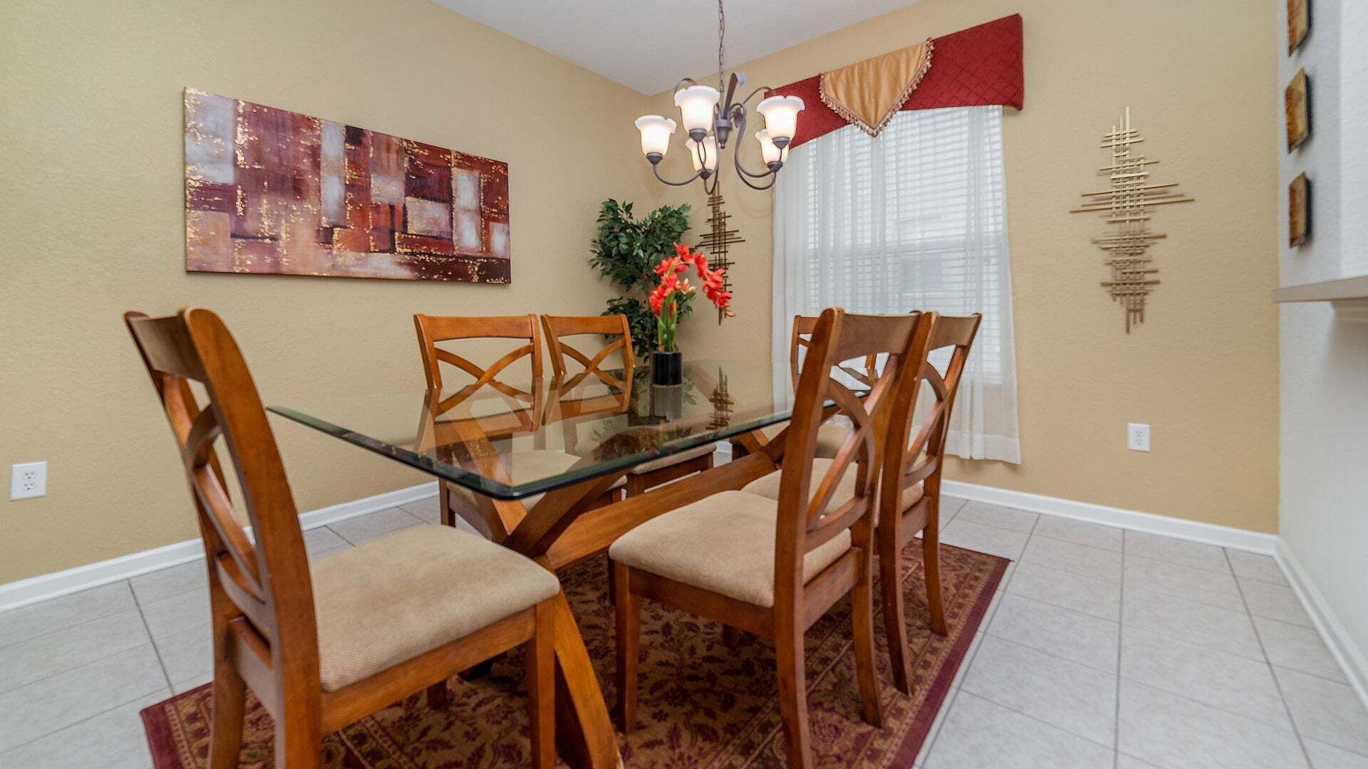 A dining room with a glass table, wooden chairs, a chandelier, a painting, a window with curtains, and a decorative wall piece.