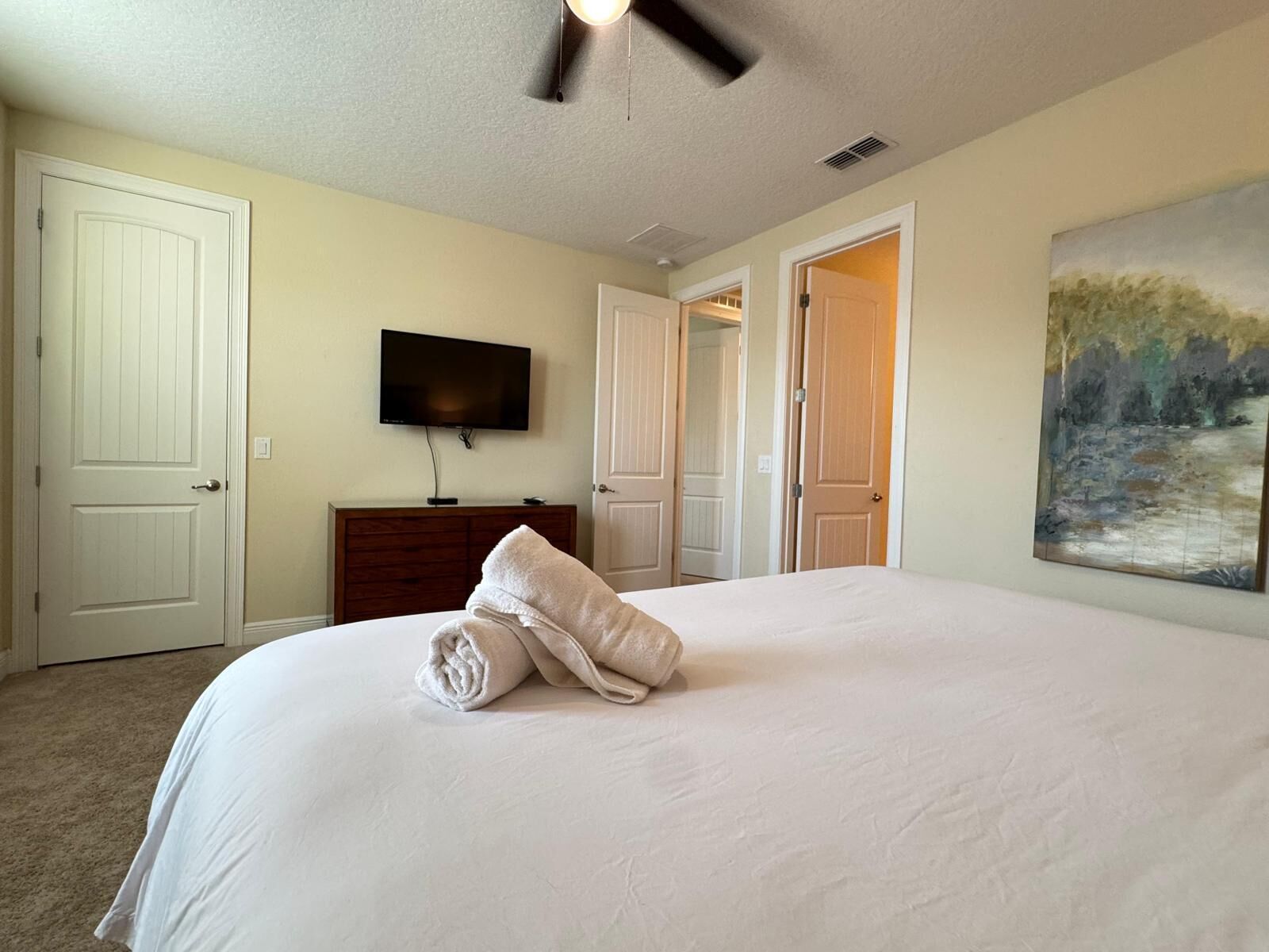 A bedroom with a large bed, a flat-screen TV mounted on the wall, a dresser, and a painting on the wall.