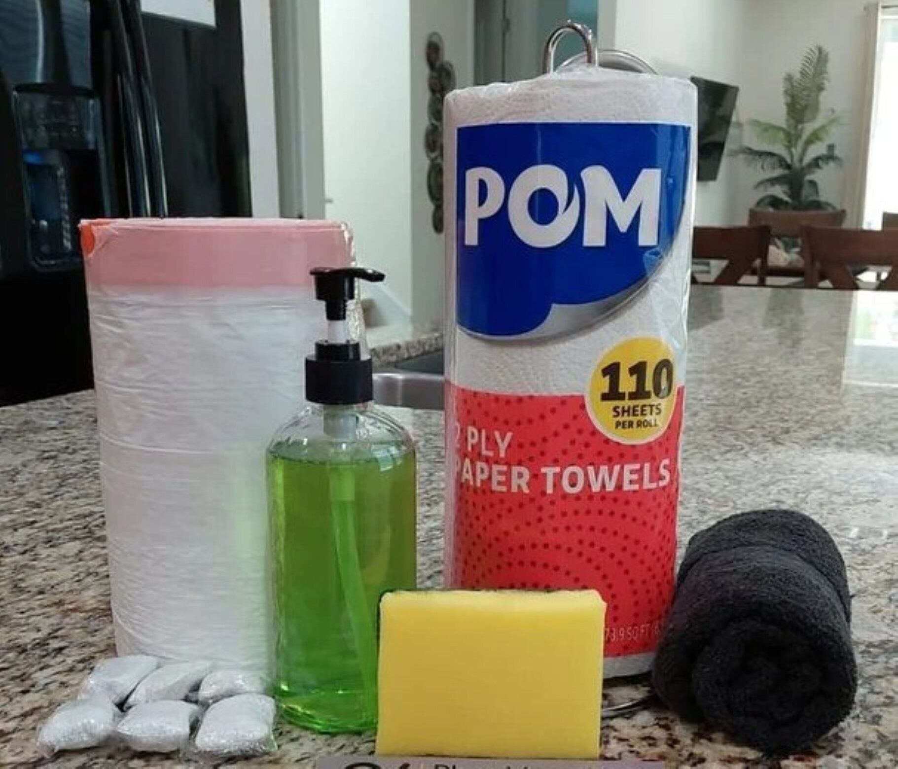 Un rouleau d’essuie-tout en papier, une bouteille de savon liquide vert, une éponge jaune et une serviette noire sur un plan de travail de cuisine.