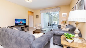 Ein modernes Wohnzimmer mit grauen Sofas, einem Couchtisch, einem Fernseher und einem Essbereich mit einer Glastür, die nach draußen führt.