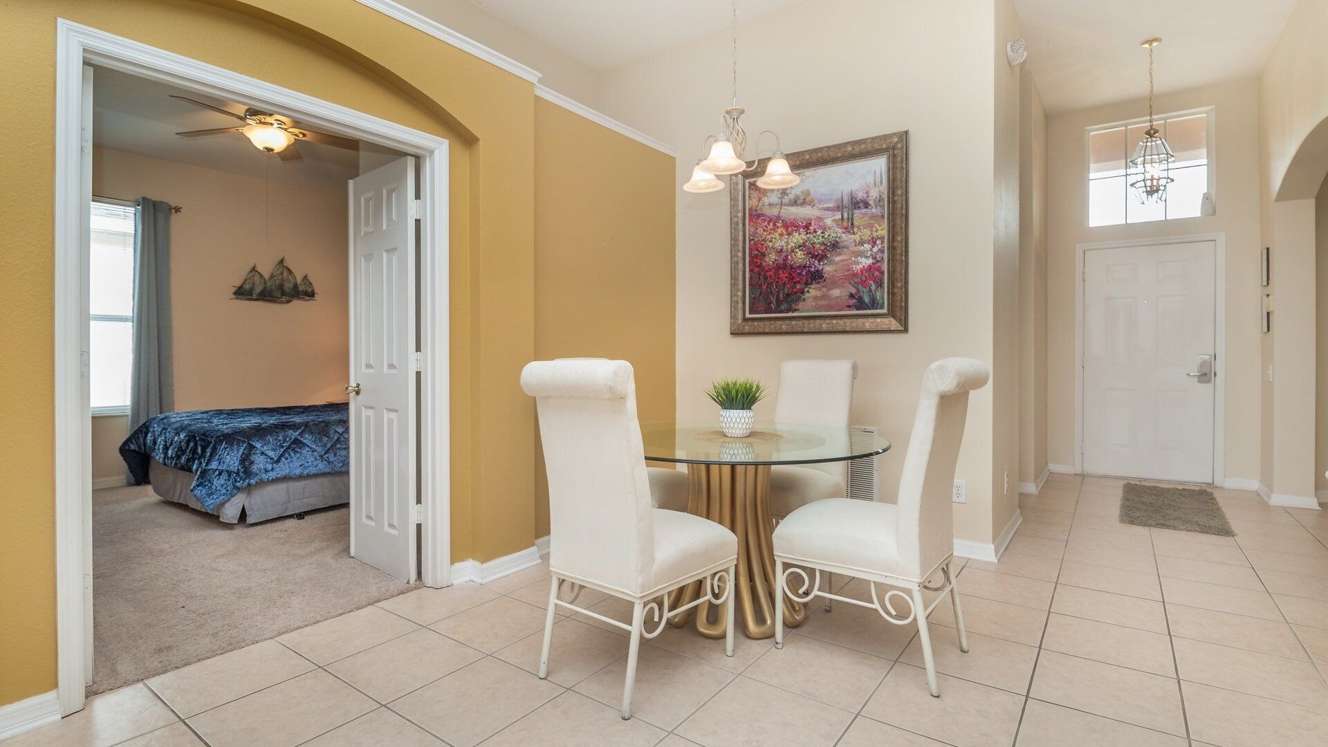 A dining area with a glass table and white chairs, a painting above the table, and a bedroom visible through an open door.