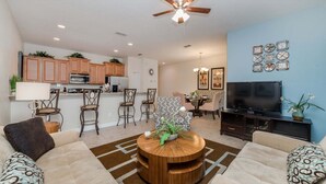A modern living room with a round wooden coffee table, a sofa, a TV, and a dining area with chairs. - Bch3031 (Kissimmee)