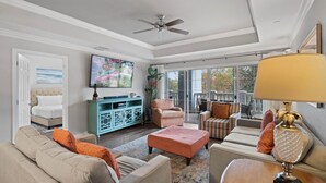 A spacious living room with a ceiling fan, a large TV, and a view of the outdoors through a sliding glass door. - Bk0203 Villas South Preserve (Kissimmee)