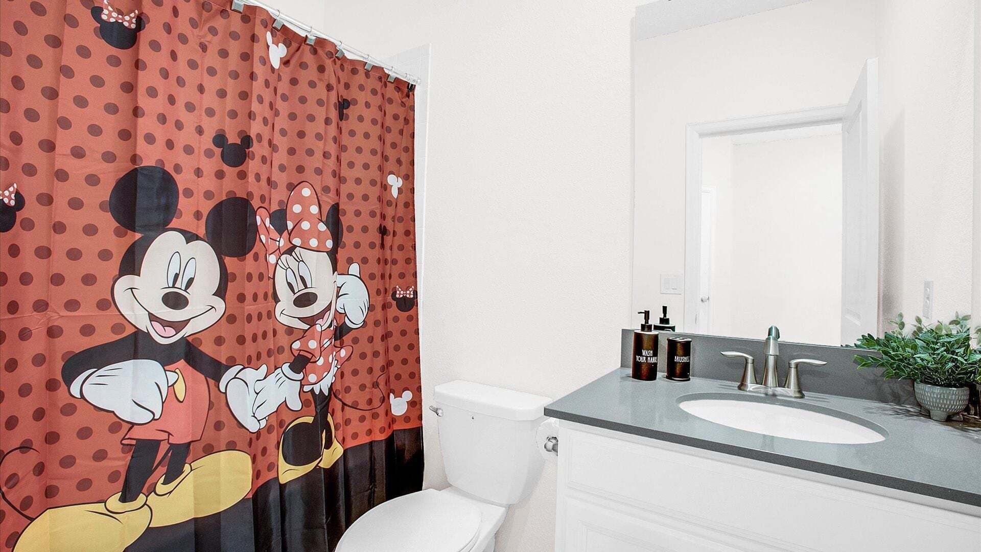 Ein Badezimmer mit Mickey Mouse Duschvorhang, Toilette, Waschbecken mit Spiegel und Toilettenartikeln.