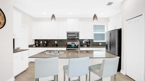 A modern kitchen with white cabinetry, a central island with chairs, a refrigerator, a microwave, and a coffee maker. - Wsb670 (Kissimmee)