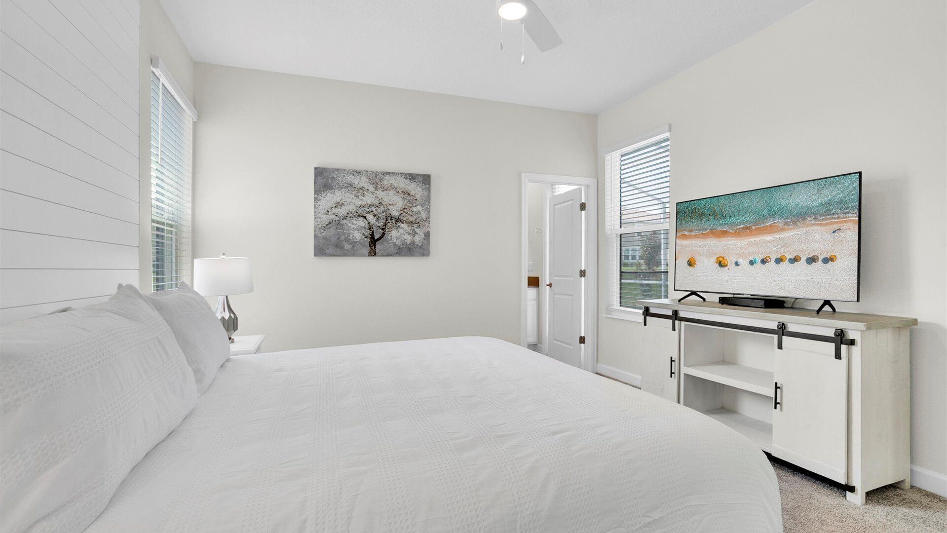 Ein modernes Schlafzimmer mit einem großen Bett, einem Fernseher auf einem weißen Schrank und einem Baumgemälde an der Wand.