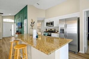 A kitchen with a granite island, wooden stools, a stainless steel refrigerator, and white cabinetry. - Pet-friendly Home w Pool Near Disney & Champions Gate (Davenport)