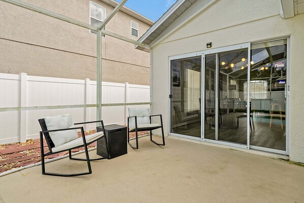 A patio with two chairs and a table, a white fence, and a sliding glass door leading to a covered area. - Pet-friendly Home w Pool Near Disney & Champions Gate (Davenport)