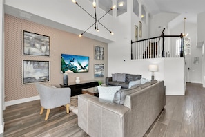 Ein modernes Wohnzimmer mit einem grauen Sofa, einem Couchtisch und einem an der Wand montierten Fernseher. Eine Treppe führt zu einer oberen Ebene.