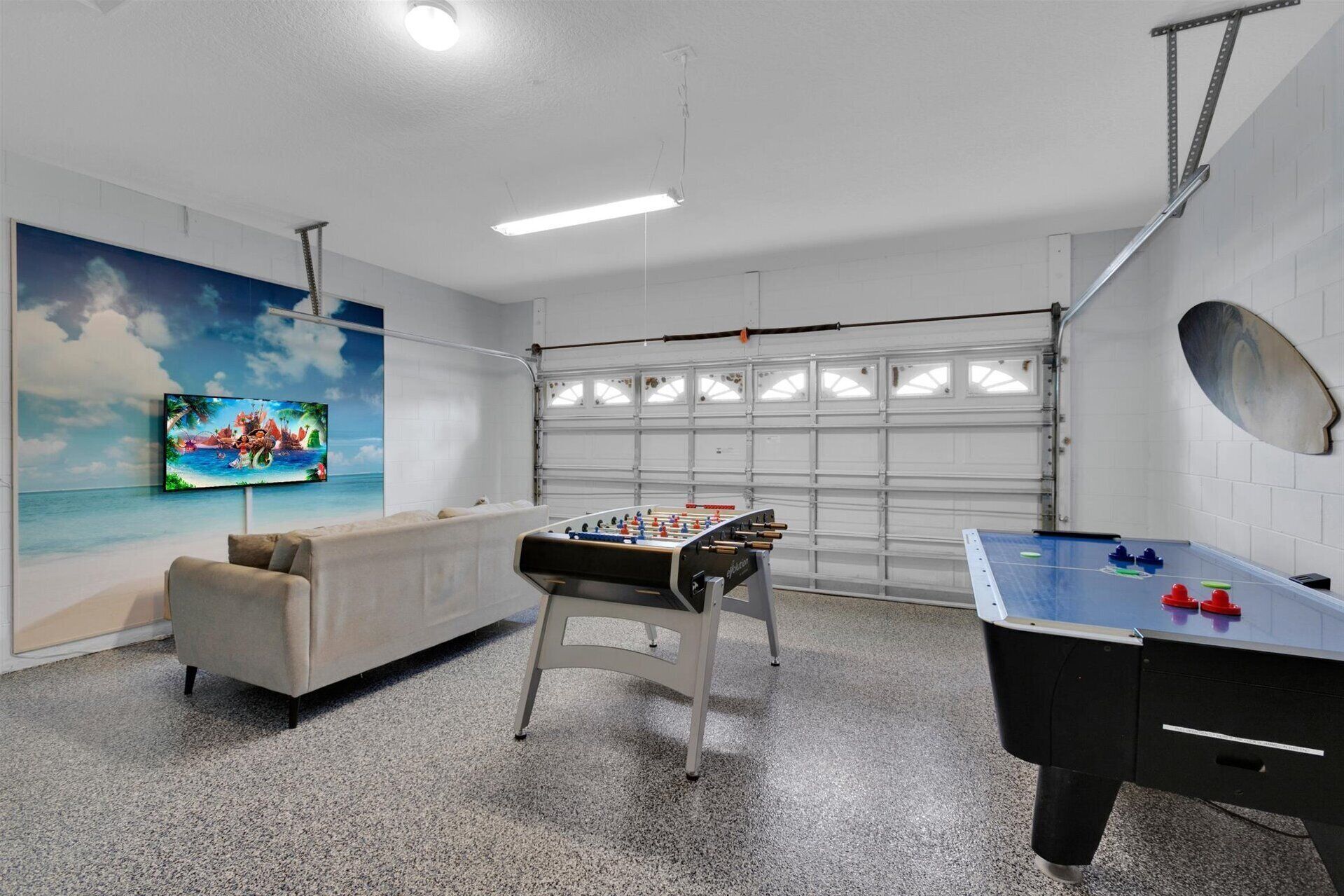 A room with a foosball table, a sofa, a TV mounted on the wall, and a garage door.