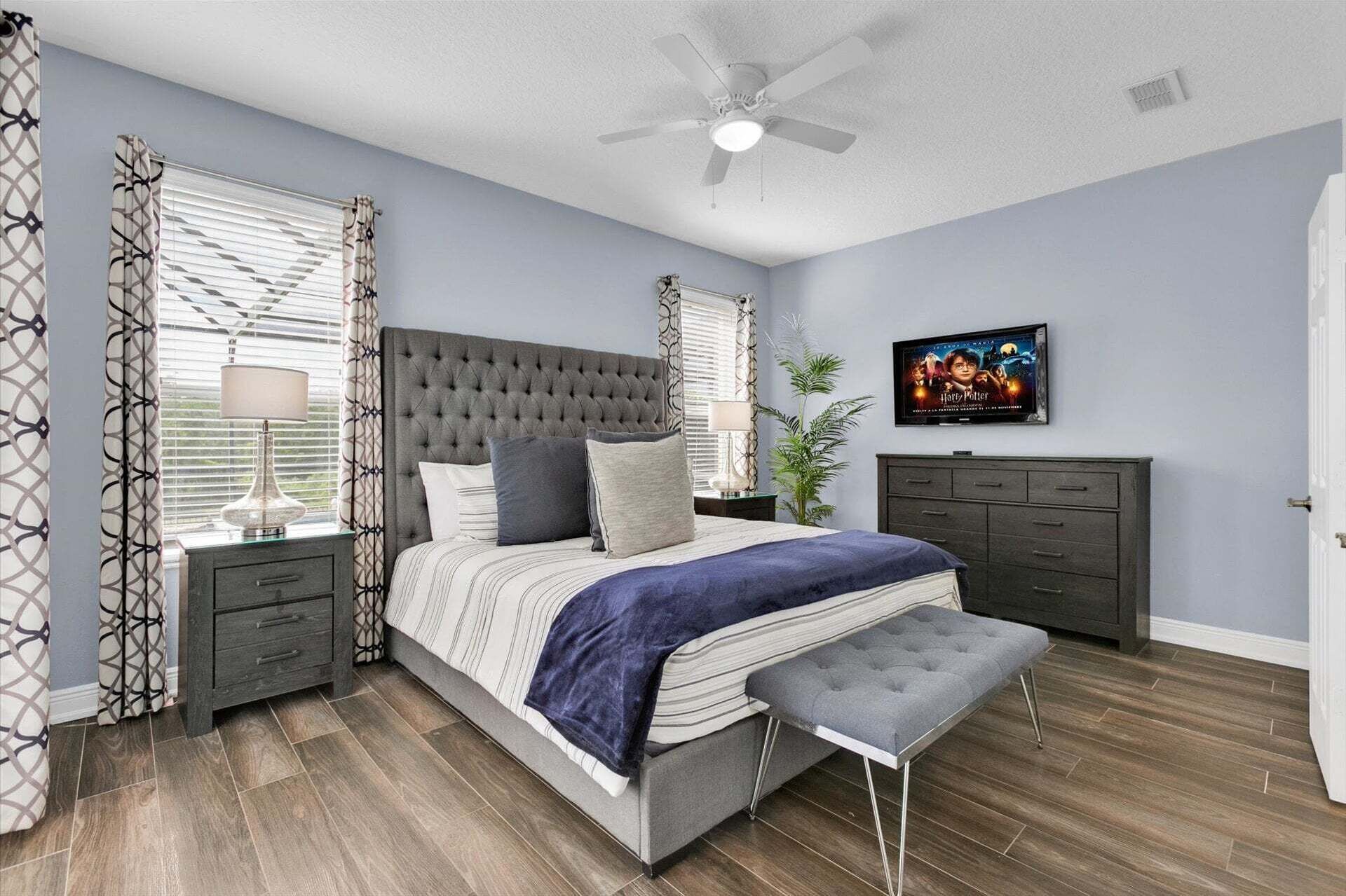Een slaapkamer met een gestoffeerd hoofdeinde, een bed met een blauwe deken, een bankje, een nachtkastje, een dressoir, een plafondventilator en een poster aan de muur.