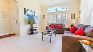 A living room with a television, a sofa with red cushions, a coffee table, and a window with blinds.