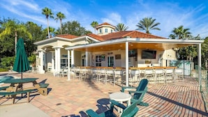 A covered outdoor area with multiple tables and chairs, a pool, and palm trees.