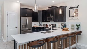A modern kitchen with dark cabinets, stainless steel appliances, a white island with bar stools, and pendant lighting. - Sts771 (Kissimmee)