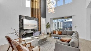 A modern living room with a large flat-screen TV, a grey sofa, wooden chairs, and a coffee table. - Ssh9191 (Davenport)