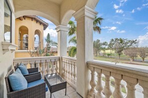 Un balcón con mobiliario de mimbre y una mesa de vidrio, con vistas a una zona ajardinada con palmeras y un edificio con arcos.