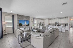 A modern living room with a sectional sofa, a flat-screen TV, and a kitchen area with white cabinets. - Solara Resort Magical Escape Private Pool & Spa (Kissimmee)