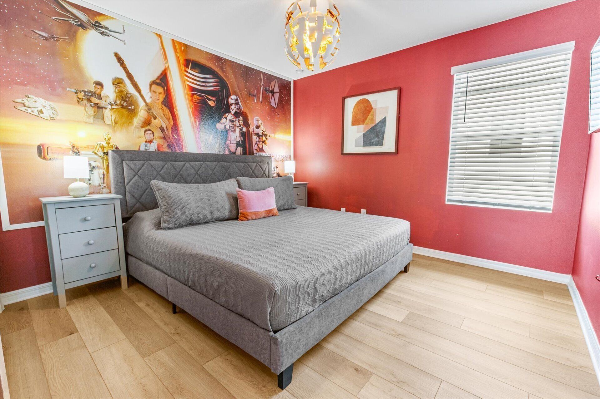 Une chambre avec un lit gris, un mur rouge, une fresque murale sur le thème de Star Wars, un lustre moderne et une fenêtre avec des stores.