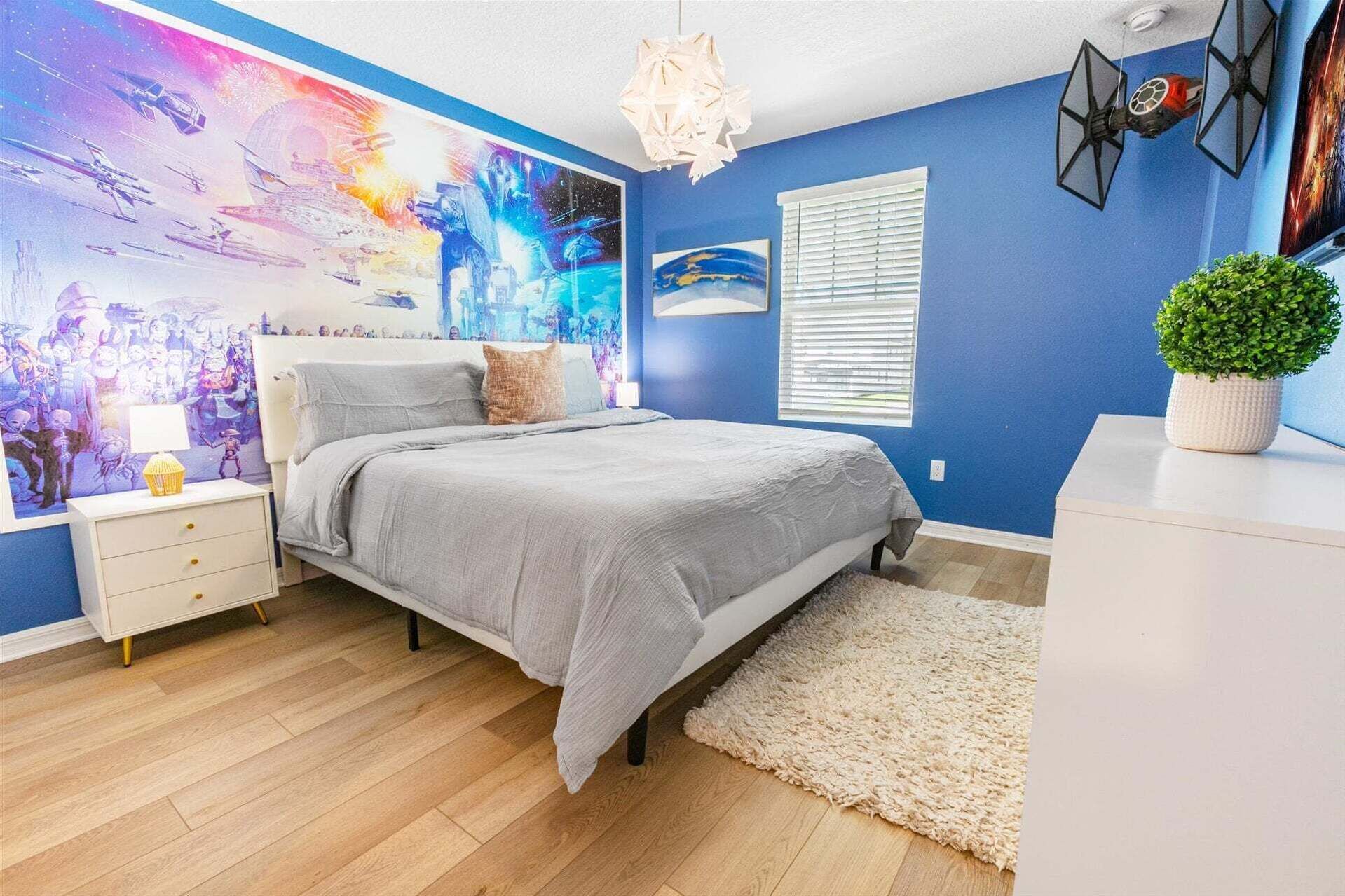 Un dormitorio con cama, mesita de noche, lámpara, decoración en la pared, ventana y una planta en maceta.