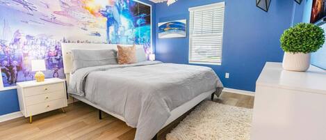 Un dormitorio con cama, mesita de noche, lámpara, decoración en la pared, ventana y una planta en maceta.