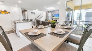 Une cuisine moderne avec des meubles blancs, un coin repas doté d’une grande table pour quatre personnes et une vue sur l’extérieur grâce à une porte-fenêtre coulissante.