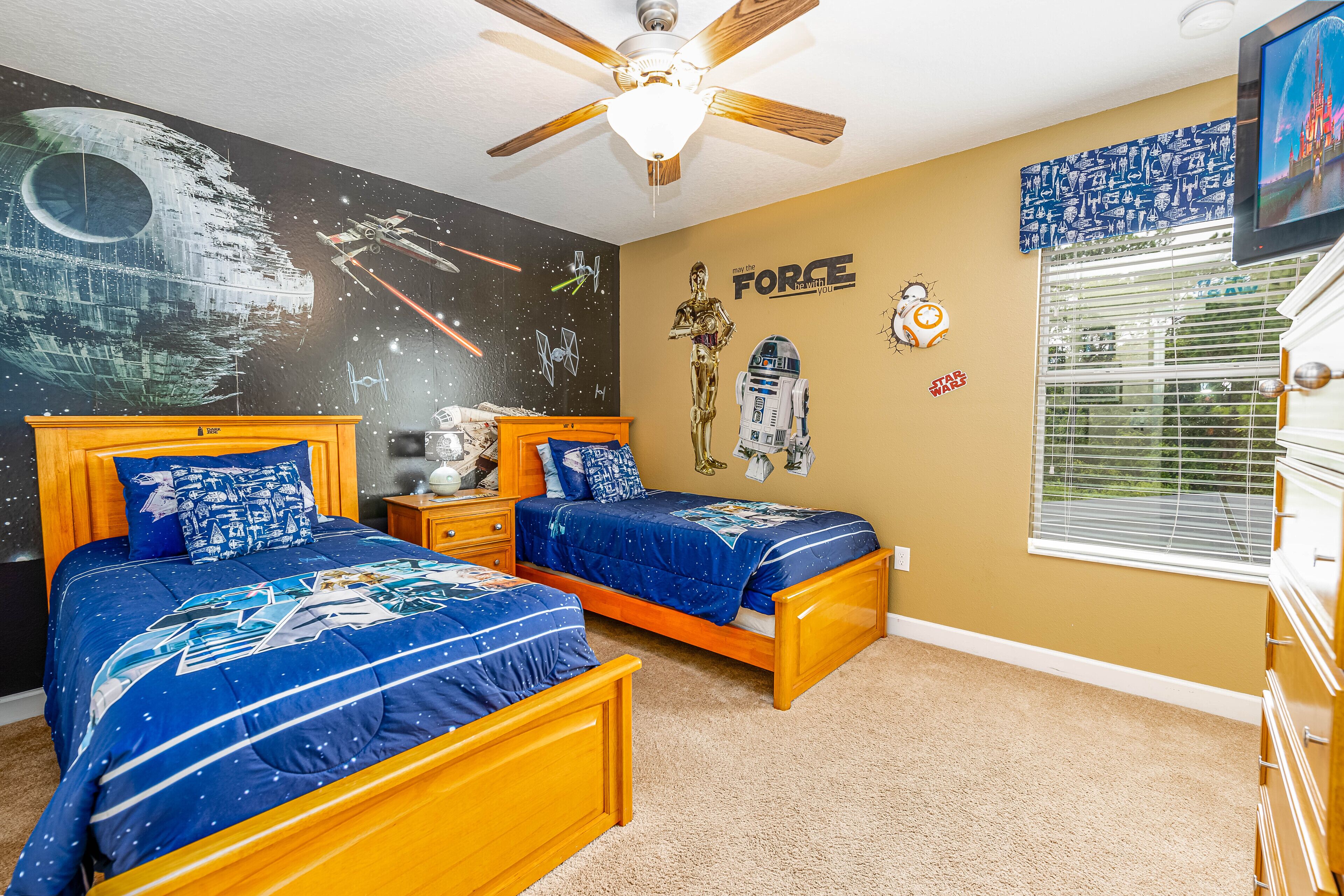A bedroom with two wooden beds, a ceiling fan, Star Wars-themed wall art, and a window with blinds.