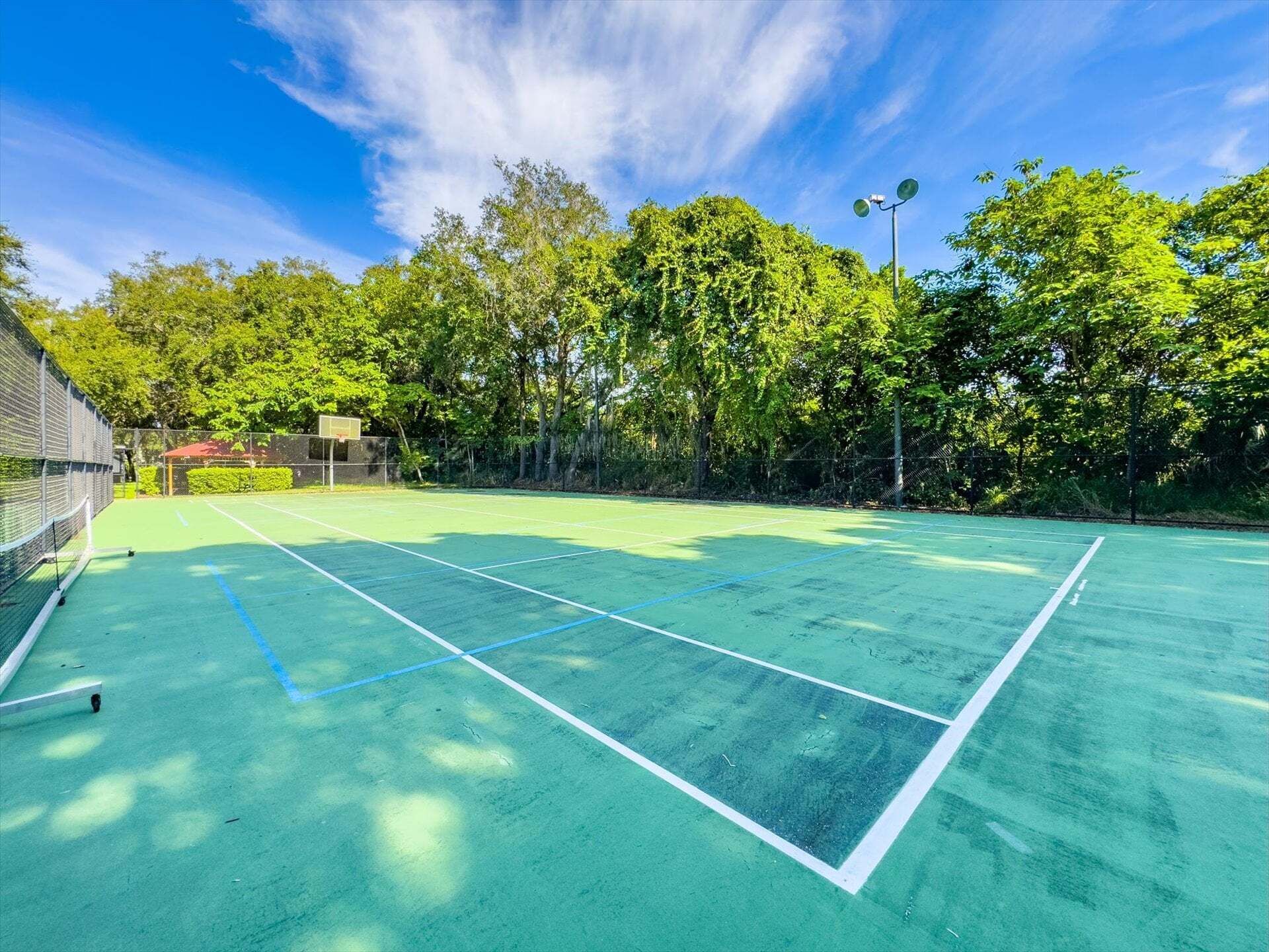 An outdoor tennis court with a net, surrounded by trees and a clear sky.