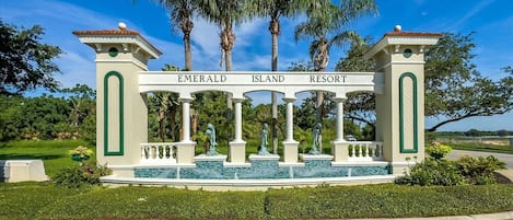 A sign for Emerald Island Resort with a classical architectural design, featuring columns and statues, surrounded by lush greenery and palm trees.