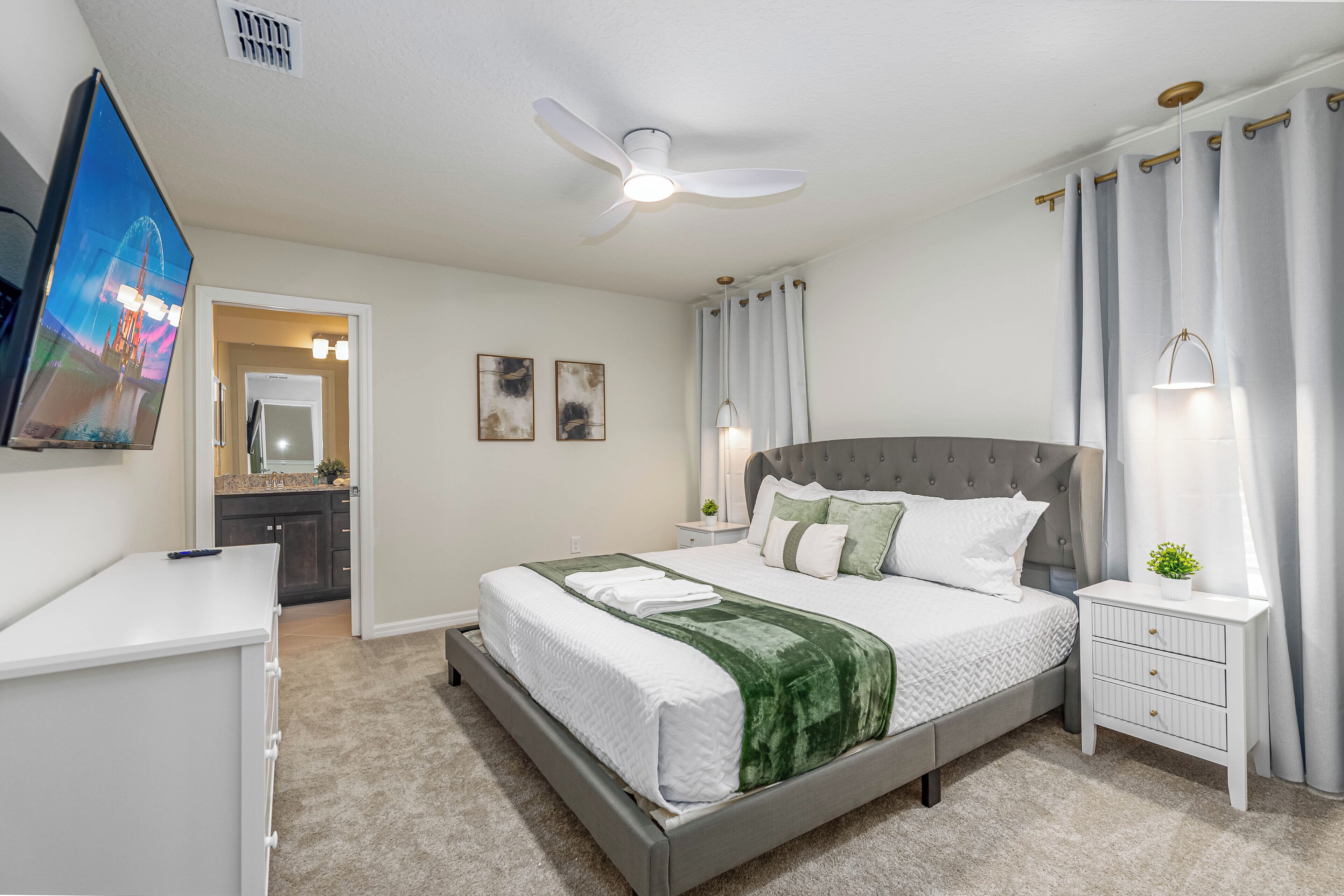 A bedroom with a bed, a nightstand, a desk, a ceiling fan, and a TV mounted on the wall.