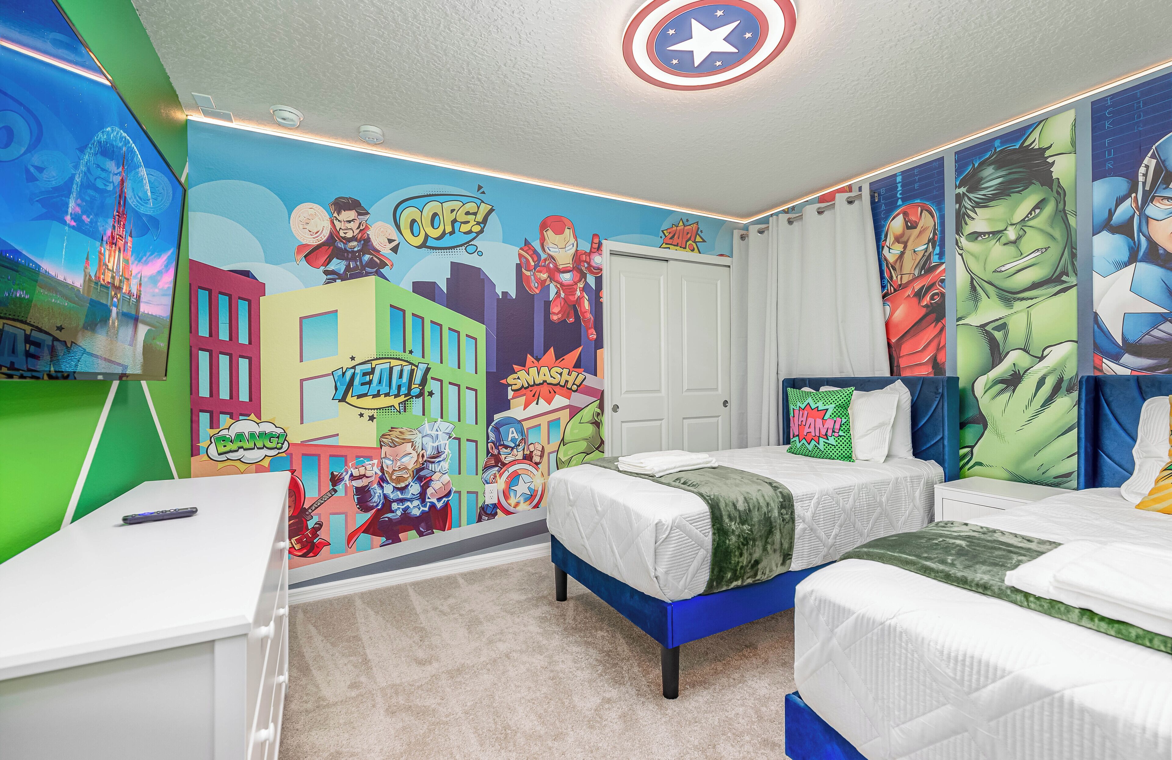 A bedroom with two beds, a desk, and a wall mural featuring superheroes.