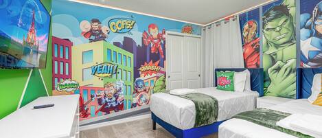 A bedroom with two beds, a desk, and a wall mural featuring superheroes.