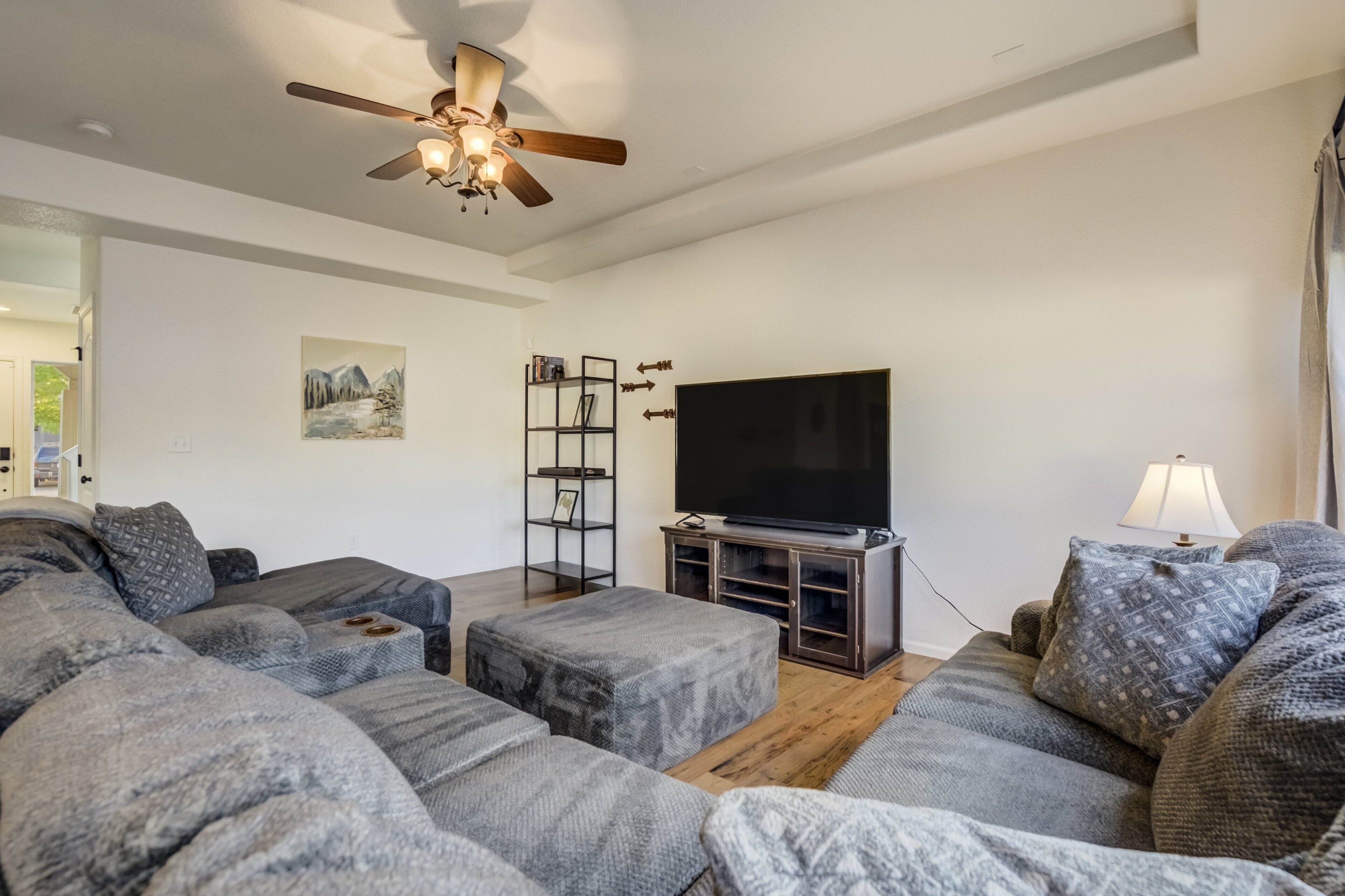 A modern living room with grey sofas, a flat-screen TV, a ceiling fan, and a bookshelf.