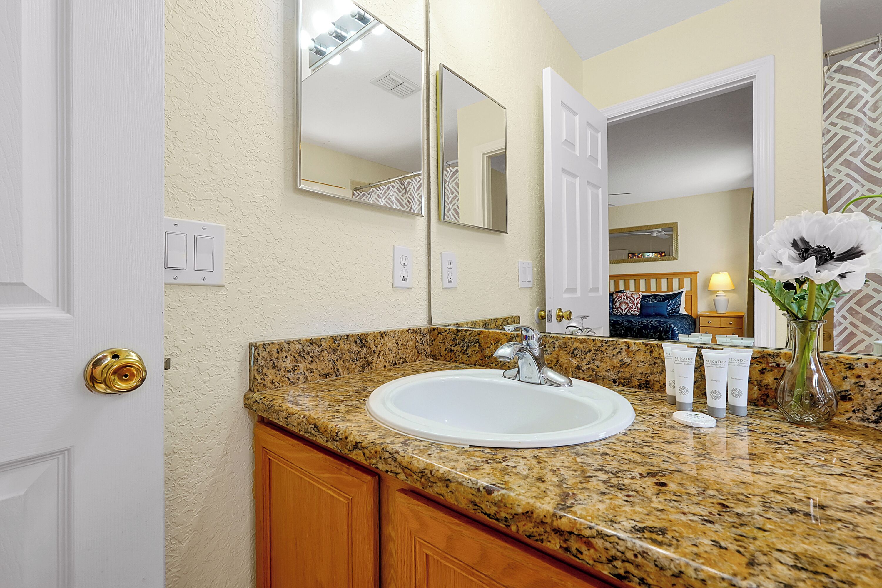 A bathroom with a granite countertop, white sink, and a mirror above it. There are toiletries on the counter and a vase with flowers.
