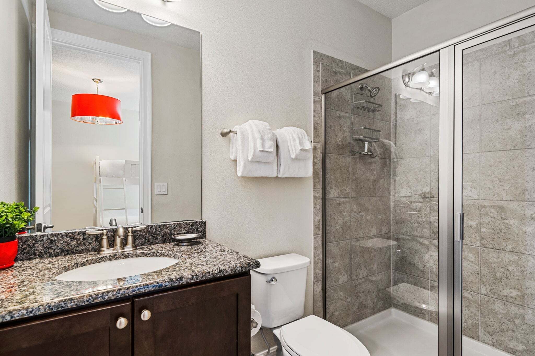 A bathroom with a double sink, a toilet, a glass shower enclosure, and a red pendant light.